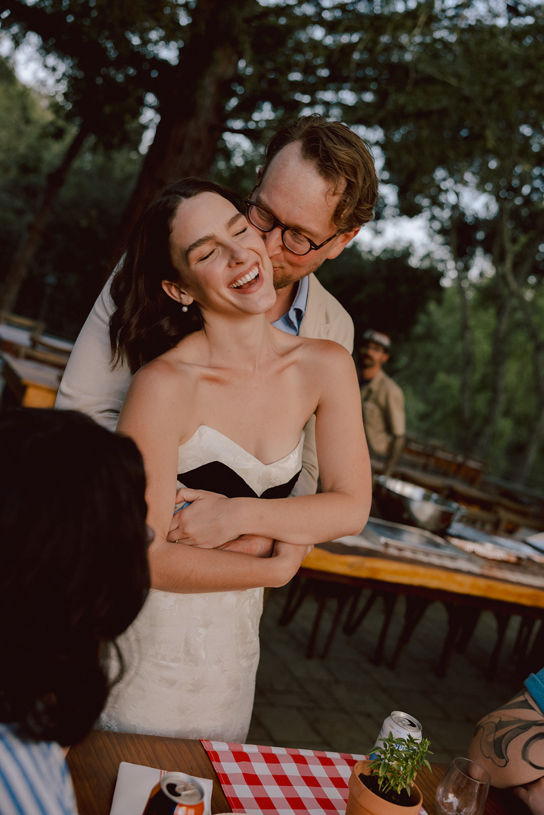 Bride and groom embracing at a reception table under the trees