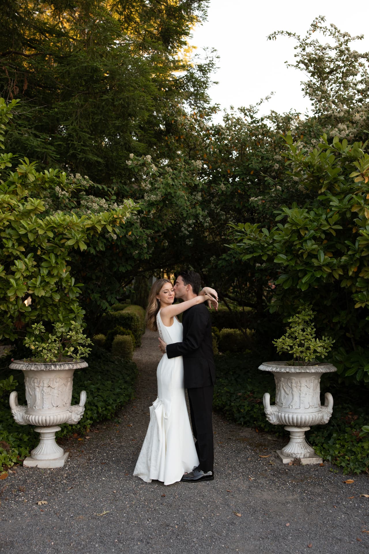 Bride and groom embracing on a gravel garden path between stone urns and hedges