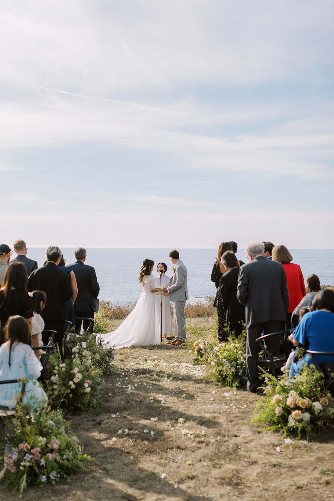 Bride and groom exchanging vows on a coastal bluff above the ocean surrounded by seated guests and wildflower aisle florals